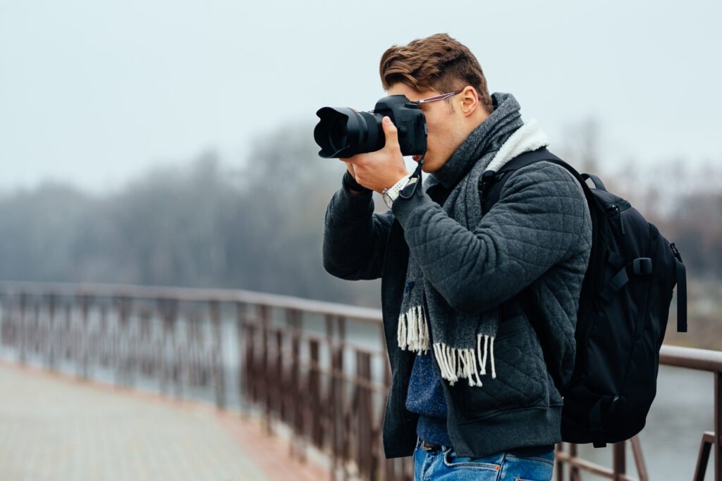 A young professional among modern photographers capturing an image with a DSLR camera while wearing a backpack and scarf outdoors