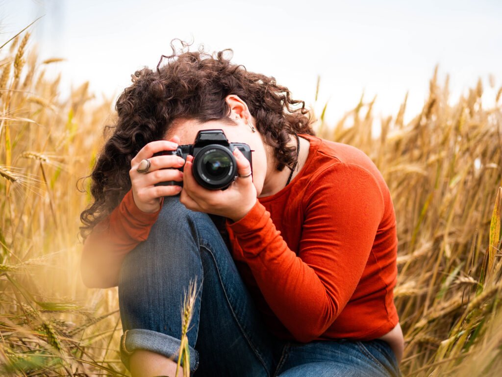 A professional affordable photographer with curly hair, wearing an orange shirt and jeans, crouched in a wheat field while focusing her camera for a creative shoot
