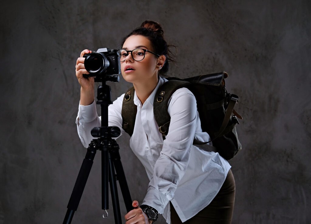 A female photographer with glasses and a backpack adjusting a DSLR camera on a tripod, representing an affordable female photographer working in a studio